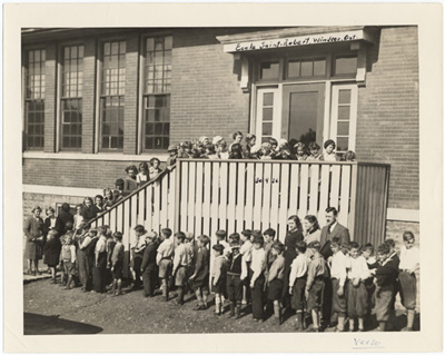 Reproduction d’une photographie de l’ouverture de la nouvelle &eacute;cole bilingue Saint-Robert, rue Wyandotte-Ouest, Windsor (Ontario), le 30 septembre 1936, l’inspecteur Robert Gauthier et les enseignantes, Mabel Parent et Fernande St-Pierre, accueillant les &eacute;l&egrave;ves, 1936.